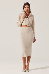 A woman is wearing a cream-colored, cropped, button-up sweater with a collar, paired with a matching high-waisted, mid-length knit skirt. She is also wearing light-colored, pointed-toe heels with ankle straps.